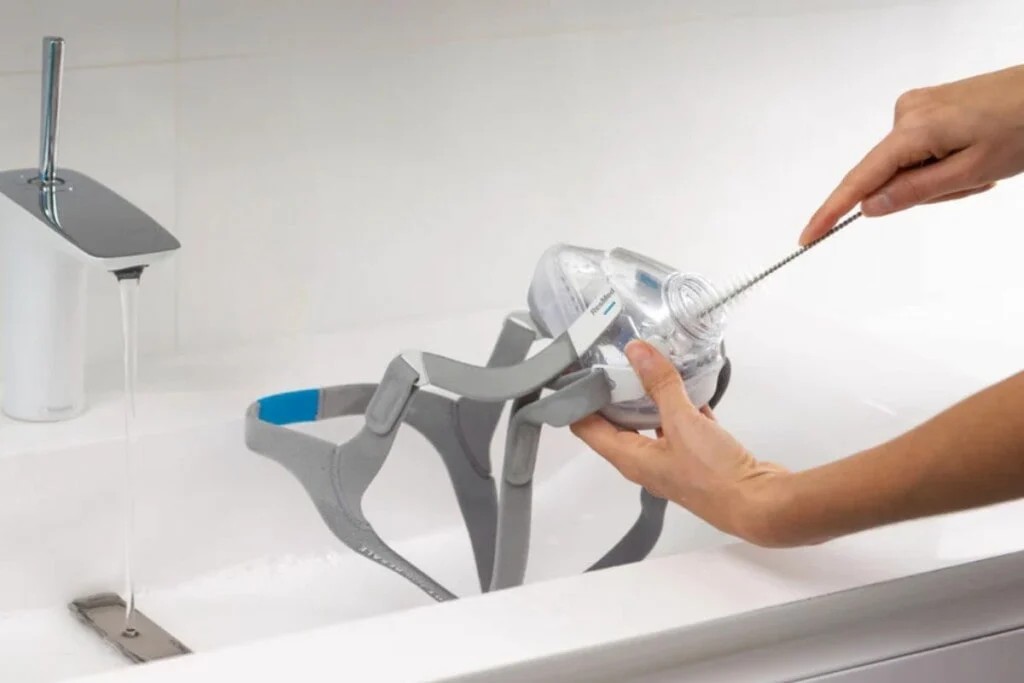 Person cleaning a CPAP full face mask with a brush at a bathroom sink